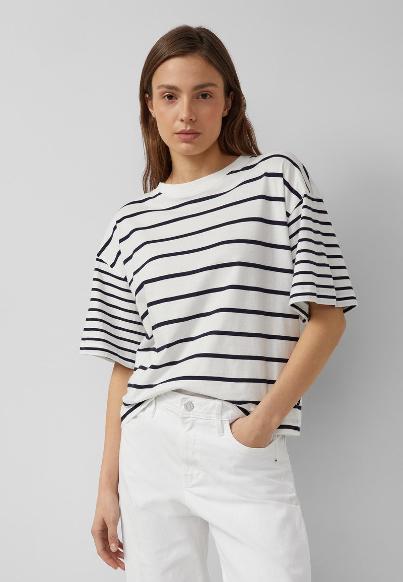 Frau, die ein weißes Kurzarmshirt mit schwarzen horizontalen Streifen und weiße Hosen trägt, posiert mit einer Hand in der Tasche vor einem grauen Hintergrund.