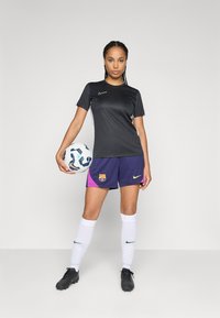 T-shirt noir à manches courtes de Nike, shorts violets et roses avec le logo de Barcelone, chaussettes blanches jusqu'aux genoux, chaussures de football noires, tenant un ballon de football à motifs.