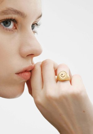 Persona joven con ojos azules apoyando la barbilla en el puño, lleva un anillo de oro con forma de corazón en el dedo medio.