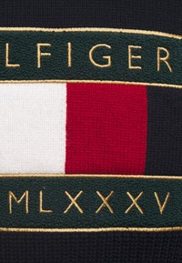 Czarna dzianina z poziomym zielonym paskiem z złotymi literami, centralnym czerwono-białym prostokątnym wzorem oraz wyszywanymi rzymskimi cyframi.