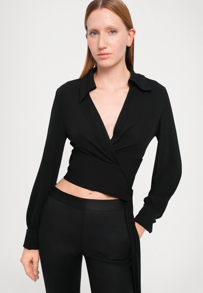 Blouse noire à manches longues avec un col structuré et un décolleté en V profond, fabriquée à partir d'un tissu lisse. Associée à un pantalon noir ajusté.