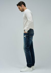 Suéter blanco texturizado para hombre, corte clásico, combinado con jeans de mezclilla de lavado medio con detalles desgastados y zapatillas blancas.