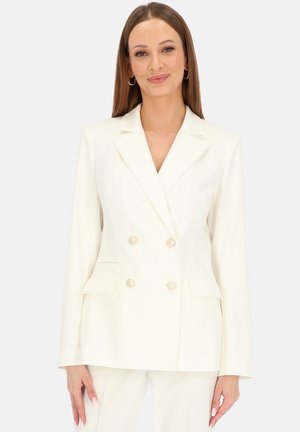 Kobieta o prostych brązowych włosach nosi biały dwurzędowy blezer z złotymi guzikami i pasujące białe spodnie, stojąc na jednolitym tle.