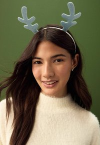 Glitrende blå gevir på en hvid hårbøjle oven på langt, mørkt hår. Modellen bærer en blød, fuzzy hvid sweater mod en grøn baggrund.