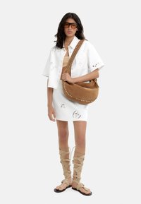 Biała koszula krótki rękaw z ażurowymi detalami, beżowy top crop, biała spódnica z wycięciami, brązowa torba o luźnym kroju oraz beżowe sandały na sznurówki.