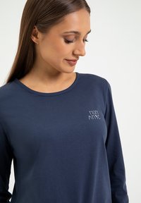 Granatowa koszulka z długim rękawem wykonana z bawełny, z okrągłym dekoltem i małym haftowanym detalem z przodu po lewej stronie. Gładka faktura.