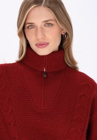 Roter Strickpullover mit hohem Kragen, Reißverschluss und strukturierten Mustern, der Kabelstrickdetails an den Seiten aufweist.