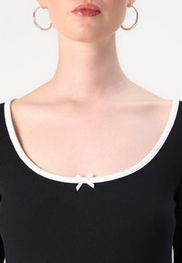 Svart ribbet topp med hvit utringning og en liten hvit sløyfe i midten. Har korte ermer og en figurnær design.