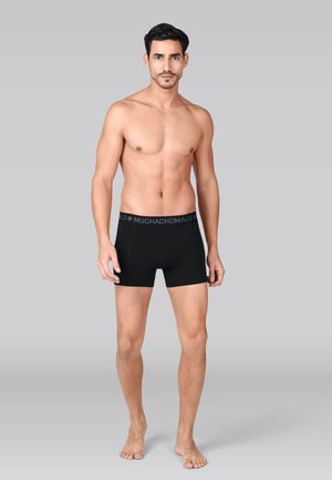 Man staat op blote voeten en draagt zwarte boxer shorts met een "MUCHACHOMALO" tailleband tegen een effen grijze achtergrond.