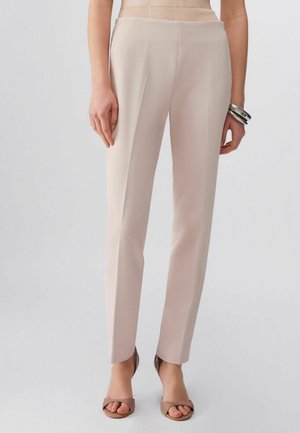 Kobieta ubrana w beżowe spodnie o prostych nogawkach i taupe sandały na obcasie z odkrytymi palcami, stojąca z luźno opuszczonymi ramionami, na prawym nadgarstku srebrne bransoletki.