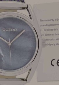 Montre argentée avec un cadran rond en bleu irisé, des aiguilles fines et un bracelet élégant. Accompagnée d'une petite étiquette de conformité.