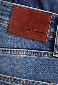 Jeans de mezclilla con un parche de cuero marrón en la cintura que presenta "Pepe Jeans London" grabado. Lavado azul claro, diseño estándar de cinco bolsillos, costuras cosidas.