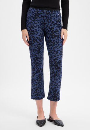 Frau trägt kurze blaue Hosen mit schwarzem Leopardenmuster und schwarze spitze Slingback-Ballerinas und steht vor einem weißen Hintergrund.