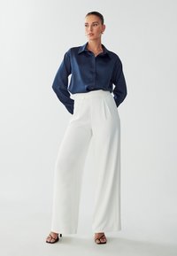 Camicia in raso blu navy con colletto, abbinata a pantaloni bianchi a vita alta. Il modello indossa sandali neri con liste. Texture lisce.