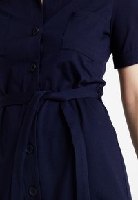 Námořnické šaty s krátkým rukávem, dvěma náprsními kapsami a zavazovacími pásky v pase. Vyrobené z měkké látky s hladkou texturou.