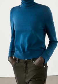 Person wearing a blue turtleneck sweater and dark brown pants with hands in pockets against a plain background.
