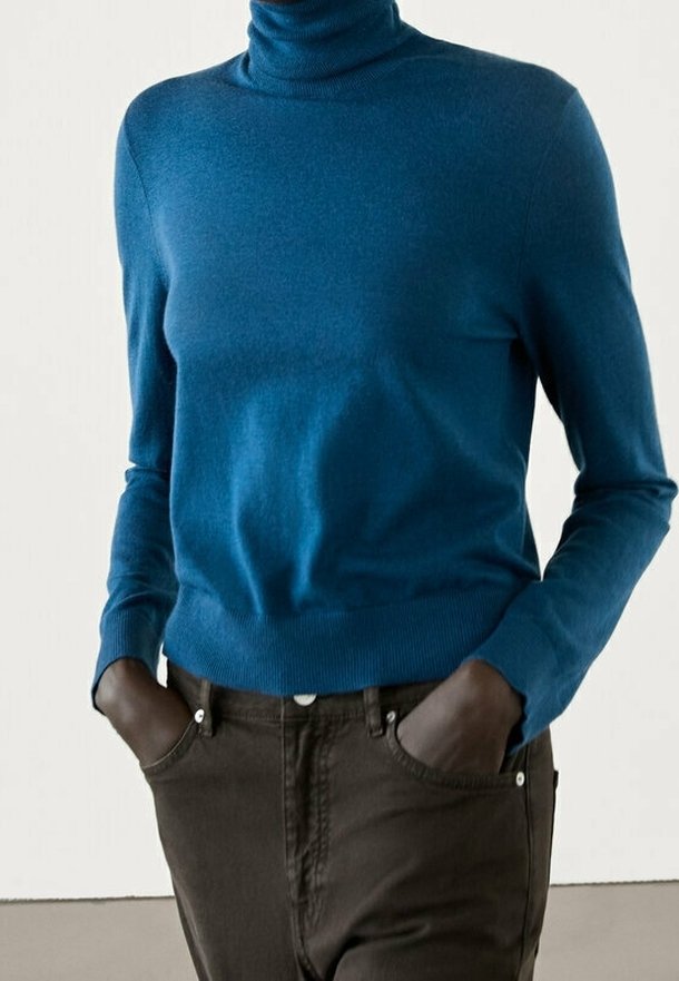 Person wearing a blue turtleneck sweater and dark brown pants with hands in pockets against a plain background.