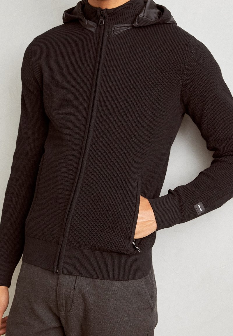 Black zip-up hoodie with ribbed texture, featuring a hood, side pockets, and a small branded tag on the cuff. Soft fabric, fitted design.