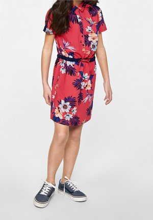 Barn iført kortærmet rød kjole med farverigt blomstermønster og marineblåt bælte, kombineret med blå sneakers med hvide snørebånd.