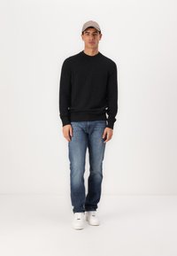 Mørkegrå strikkegenser, blå denimjeans og hvite joggesko. Modell iført en lysegrå caps, stående mot en ensfarget hvit bakgrunn.