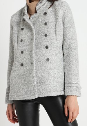 Grå uld dobbeltradet frakke med dekorative sorte knapper båret over en beige rullekrave og sorte læderbukser.