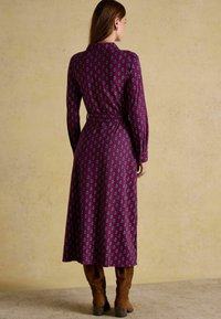 Fialové šaty s dlhými rukávmi a geometrickým vzorom, s opaskom v páse a splývavým strihom do polovice stehien. Nosené s hnedými čižmami.
