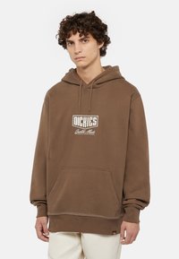 Brun huvtröja gjord av mjukt material med en framficka, som har en vit logo och sömmar vid nederkant och ärmar.