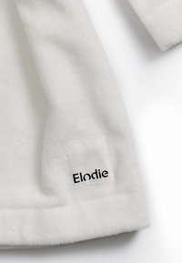 Vit tygplagg med mjuk textur, med en synlig sydd etikett som läser "Elodie" i svart på en fyrkantig lapp.