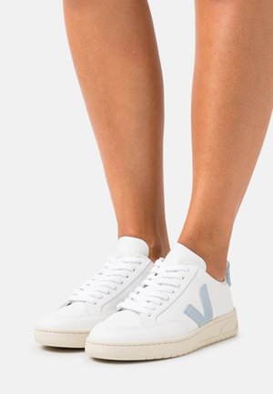 Ben klædt i hvide lædersneakers med lyseblå "V" logoer, hvide snørebånd og beige såler, vist mod en ensfarvet lys baggrund.