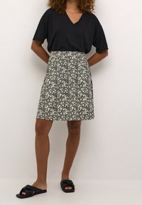 Donna che indossa una T-shirt nera a V, una gonna floreale bianca e nera e delle ciabatte nere, in piedi contro un muro bianco uniforme.
