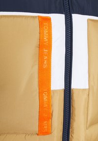 Framifrån av en beige, vit och marinblå jacka med en svart dragkedja och en orange tygskylt med texten "Tommy Jeans."