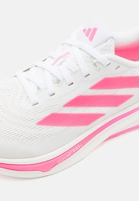 Chaussure de sport blanche avec une tige en mesh texturé, accents roses et trois rayures roses. Elle possède un laçage traditionnel et un marquage audacieux.