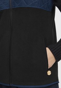 Veste en polaire noire avec une section supérieure matelassée bleu marine, dotée d'une fermeture éclair et de poches latérales. Accent rond en bois près de l'ourlet.