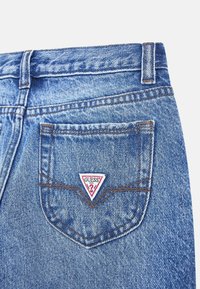 Des jeans en denim avec un délavage bleu clair comportent une poche arrière avec un logo triangulaire "GUESS" blanc et rouge et des coutures marron contrastantes.