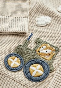 Beige stickad cardigan med texturerad tyg, ribbade manschetter och en grön traktor broderad med ett leende ansikte och dekorativa hjul.