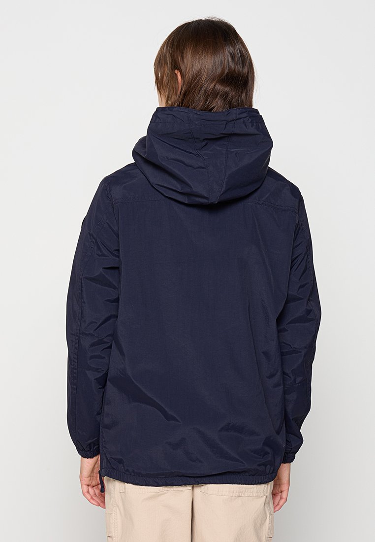 Personne aux cheveux bruns de longueur moyenne portant une veste à capuche bleu marine et un pantalon beige, de dos contre un fond clair.