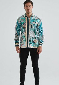 Homme debout portant une chemise à manches longues avec des motifs architecturaux et floraux multicolores et un pantalon noir.