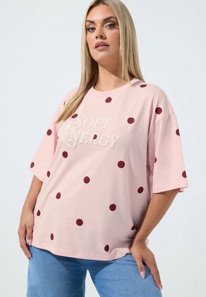 Frau mit langen blonden Haaren trägt ein pinkes übergroßes T-Shirt mit dunkelroten Punkten und dem Text "SOFT ENERGY", kombiniert mit blauen Jeans.