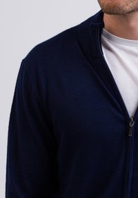 Granatowy sweter zapinany na zamek o wysokim kołnierzu, z prążkowaną teksturą i widocznymi srebrnymi elementami zamka, nałożony na białą koszulę.