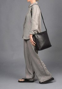 Bolso de hombro de cuero negro con correa ajustable; textura suave y diseño minimalista. Combinado con una camisa de lino gris claro y pantalones anchos.