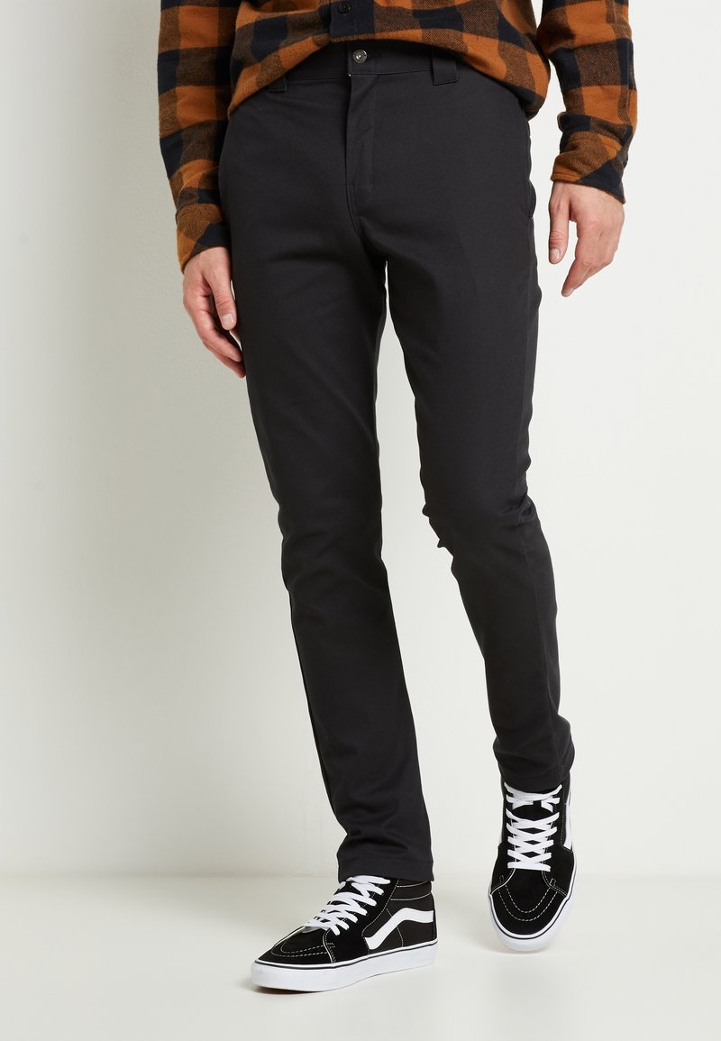 Mustat slim-fit-housut, jotka on valmistettu sileästä kankaasta, klassisella leikauksella ja sivutaskuilla, yhdistettynä mustiin ja valkoisiin high-top-tennareihin.