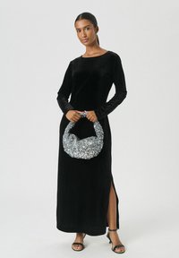 Femeie îmbrăcată într-o rochie lunga din catifea neagră, cu o fanta pe lateral, ținând o geantă din paiete silver și purtând sandale negre cu vârf deschis pe un fundal alb.