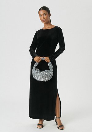 Frau in einem langen schwarzen Samtkleid mit seitlichem Schlitz, die eine silberne Paillettentasche hält und schwarze offene High Heels trägt, vor einem weißen Hintergrund.