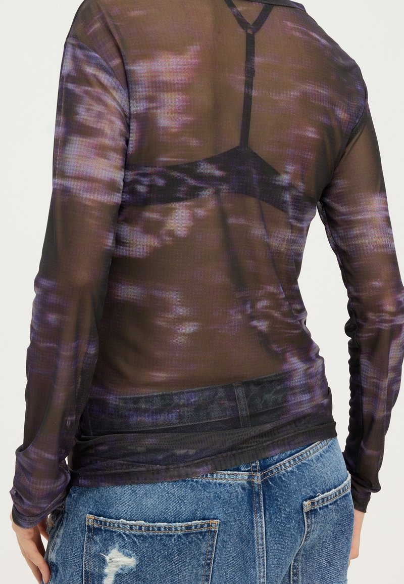 Personne portant un top transparent à manches longues avec un motif tie-dye violet, laissant apparaître un soutien-gorge noir en dessous, associé à un jean bleu déchiré.