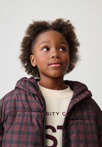 Doudoune à carreaux bordeaux et noirs avec capuche, dotée d'une fermeture éclair frontale. Chemise beige à col rond avec texte rouge en dessous.