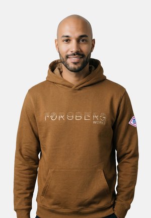 Uomo con barba curata che indossa una felpa con cappuccio marrone FORSBERG WORKS con tasca frontale e patch sulla manica sinistra, sorridendo su uno sfondo semplice.