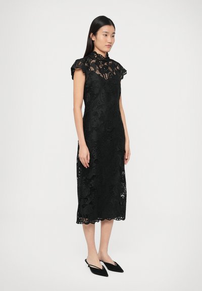 Kobieta ubrana w czarną koronkową sukienkę midi z krótkimi rękawami i wysokim kołnierzem, w połączeniu z czarnymi, spiczastymi butami na obcasie typu slingback, stojąca na białym tle.