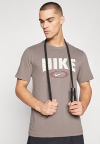Grå t-skjorte med korte ermer som har en strukturert hvit "NIKE"-logo og en bordeauxfarget oval. Modellen holder et svart hoppetau.