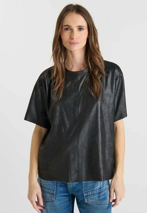 Femme aux longs cheveux bruns portant une chemise noire en simili cuir à manches courtes et un jean bleu, devant un fond uni.