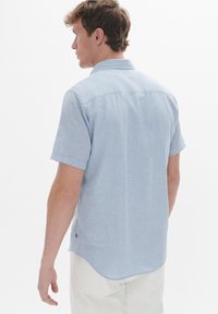 Camicia azzurra a maniche corte con colletto a bottoni. Presenta un tessuto testurizzato, un orlo curvo e un piccolo dettaglio del logo sul lato.